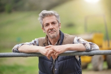 farmer facing camera and leaning on a fence, with sunlight in background